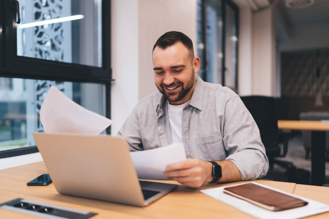 Positive male entrepreneur sitting at wooden desk at workplace going through papers while working online on business plan on laptop