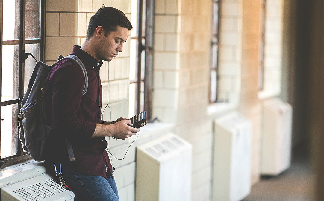 Adult student with backpack and earbuds in leaning against a wall in a hallway